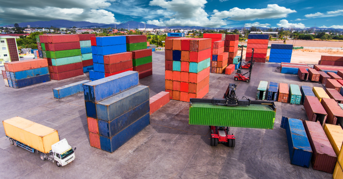 Aerial view of a bustling shipping yard with stacked colorful shipping containers. A crane lifts a green container. Truck nearby under a partly cloudy sky.