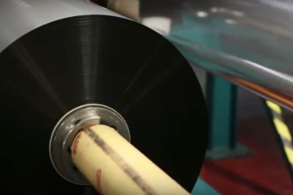 Close-up of a large black roll of film on a spool in an industrial setting. The machine parts are metallic, conveying a sense of manufacturing and efficiency.