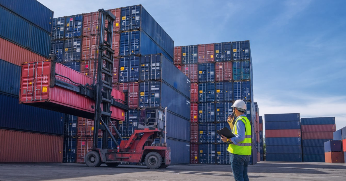 A worker in a hard hat and safety vest observes a forklift moving a red shipping container in a busy port, surrounded by stacked containers under a blue sky.