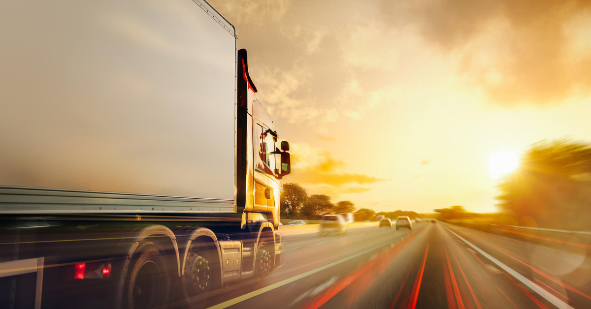 A large truck speeds along a highway at sunset, with a blurred motion effect. It conveys motion and urgency, set against a vibrant orange sky.